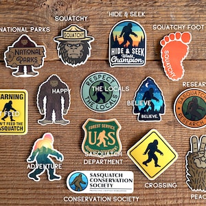 May include: A collection of 14 colourful stickers featuring a cartoon Sasquatch in various poses and settings. The stickers include phrases like "Squatchy", "Hide & Seek World Champion", "Respect the Locals", "Believe", "Sasquatch Research Team", "Warning Don't Feed the Sasquatch", "Adventure", "Sasquatch Conservation Society", "Crossing", and "Peace".