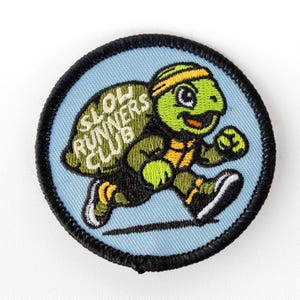 May include: A circular embroidered patch with a light blue background and a black stitched border. It features a green turtle wearing a yellow headband and running shoes, with the text SLOW RUNNERS CLUB embroidered in white on its shell.