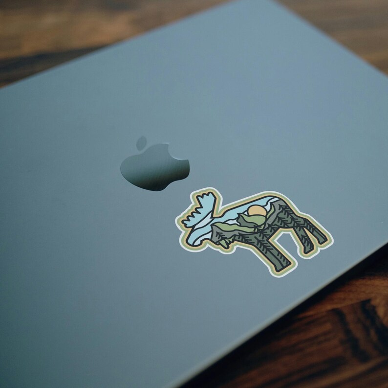 May include: A sticker featuring a moose silhouette with a landscape design inside. The design includes mountains, trees, and a sun. The sticker is placed on a gray laptop.