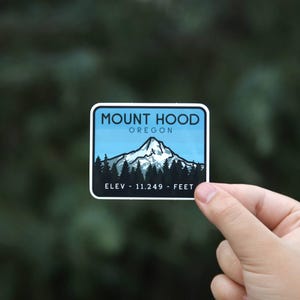 A hand holds the Mount Hood Oregon sticker against blurred dark evergreens, snowy peak and treeline in view.