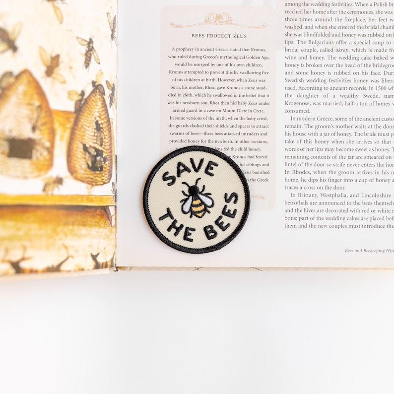 The Save The Bees patch sits on an open book page about bees protecting Zeus, its bee stitched in gold and black.