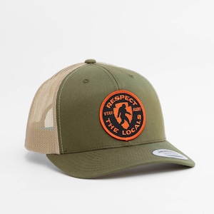 May include: An olive green trucker hat with a tan mesh back and a circular patch on the front. The patch features an orange border, black background, and an orange silhouette of a creature with the text "RESPECT THE LOCALS" and "STAY ALERT".