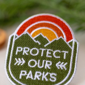 Close crop on the Protect Our Parks patch beside a tan leather patch and pine sprigs.