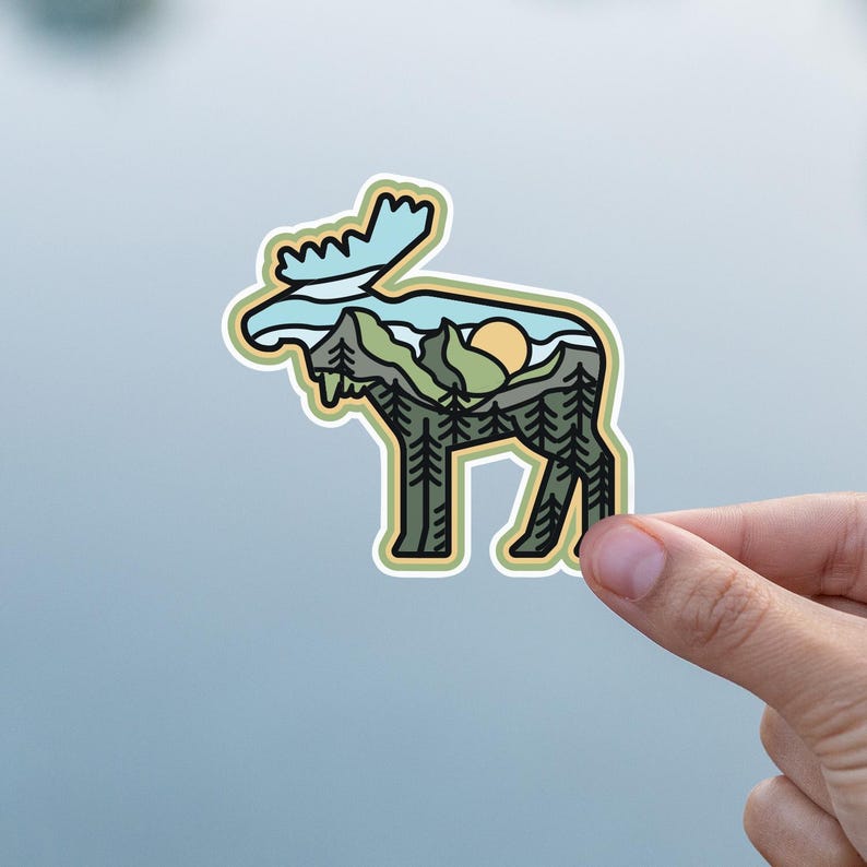May include: A die-cut sticker featuring a moose silhouette with a landscape design. The interior design includes a mountain range, trees, and a setting sun, with a light blue sky. The sticker has a yellow border and is held by a hand.