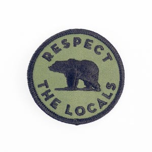 May include: A circular embroidered patch featuring an olive green background with a dark charcoal bear silhouette in the centre. The text "RESPECT THE LOCALS" is stitched in dark charcoal thread, curving along the top and bottom edges.