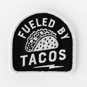 May include: A black embroidered patch with a white border, featuring a white taco graphic in the center. The text FUELED BY is arched above the taco, and the word TACOS is written below, followed by a white lightning bolt graphic.
