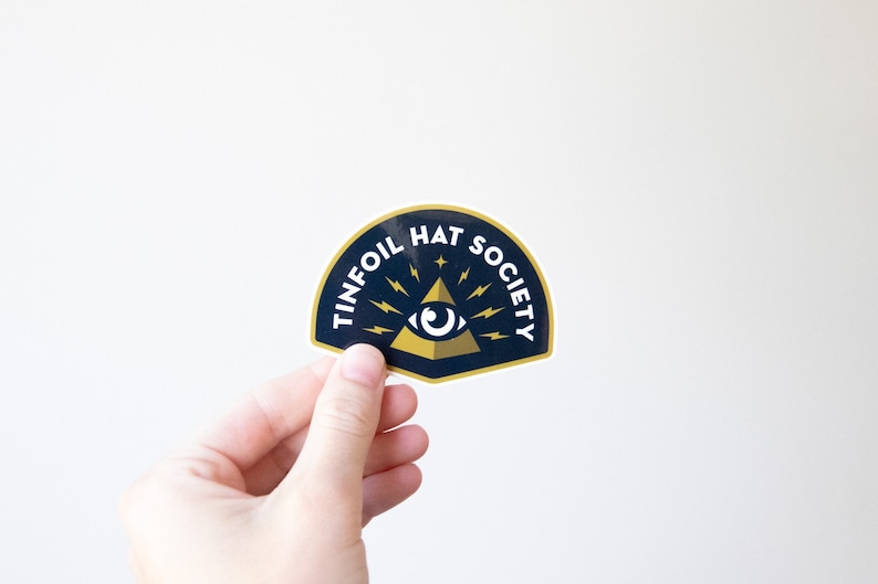 A hand holds the Tinfoil Hat Society sticker against a plain white wall, its gold eye and pyramid showing.