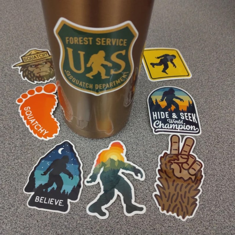 May include: A collection of nine stickers featuring a Bigfoot theme. The stickers are on a brown metal water bottle and a grey surface. The stickers feature a variety of designs, including a Bigfoot footprint, a Bigfoot with a peace sign, a Bigfoot with a sunset, and a Bigfoot with a forest scene. The stickers also feature the text "Squatchy", "Believe", "Hide & Seek World Champion", and "Forest Service US Sasquatch Department".