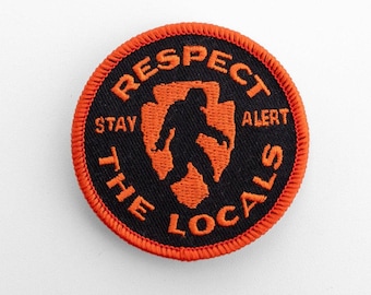 May include: A circular embroidered patch with a black background and a bright orange border. The design features an orange silhouette of a creature resembling Bigfoot centered within an orange arrowhead shape, with the text RESPECT THE LOCALS.
