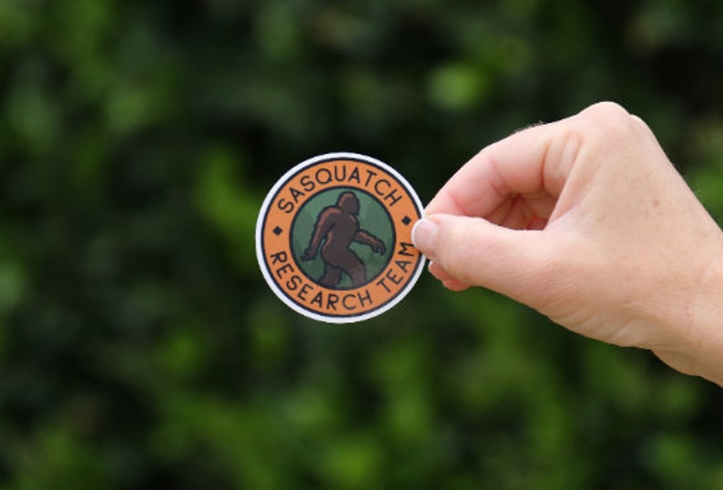 May include: A round sticker with an orange border and a green centre. The sticker features a silhouette of a sasquatch walking and the text "Sasquatch Research Team".