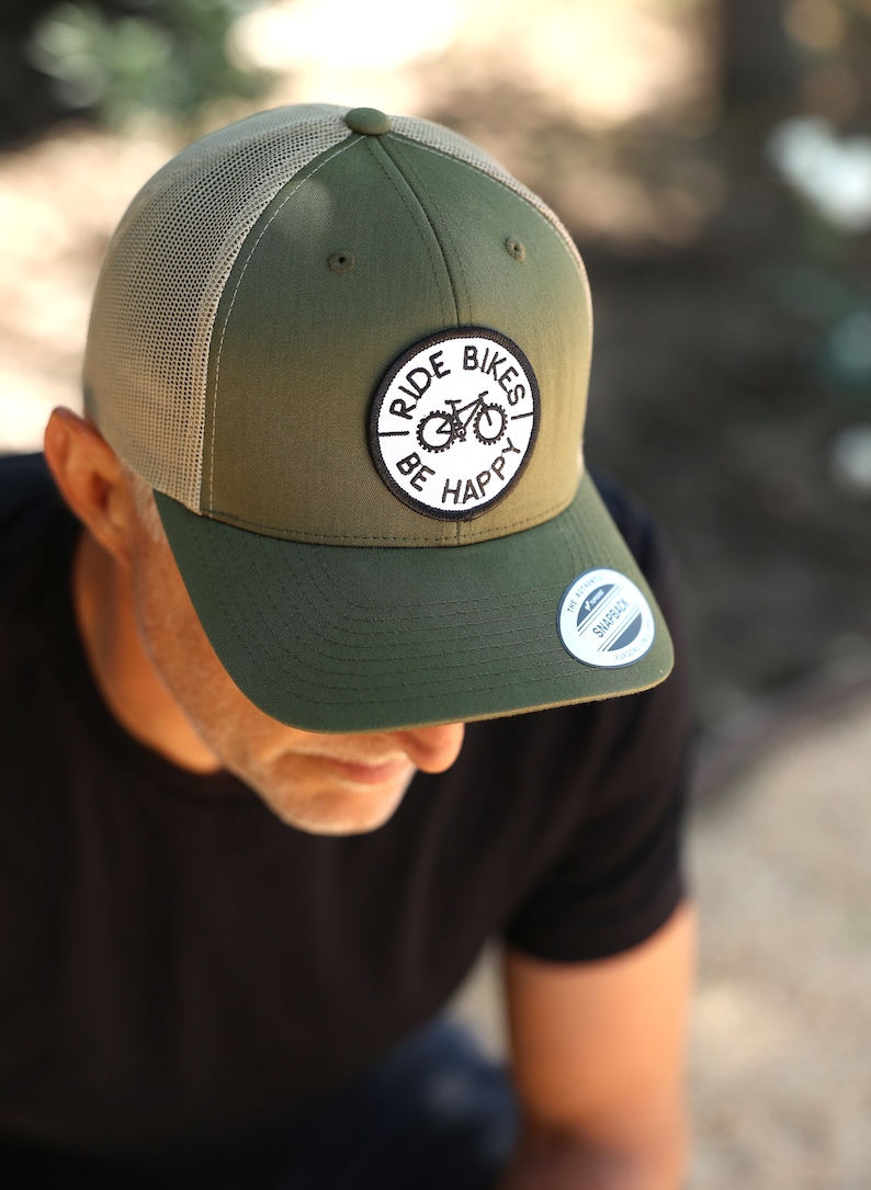 May include: A green and tan trucker hat with a black and white patch that says "Ride Bikes Be Happy" and a black and white sticker that says "The Original Snapback".