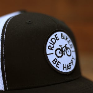 May include: Black and white trucker hat with a white mesh back. The hat has a circular patch with the text "Ride Bikes Be Happy" and a bicycle graphic.