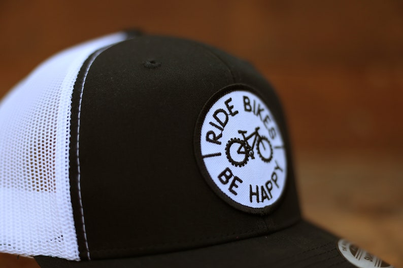 May include: Black and white trucker hat with a white mesh back. The hat has a circular patch with the text "Ride Bikes Be Happy" and a bicycle graphic.