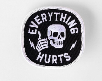 May include: A square-shaped embroidered patch with rounded corners featuring a black background and a white border. The design includes a white skull giving a thumbs up, two small lightning bolts, and the text EVERYTHING HURTS in white.