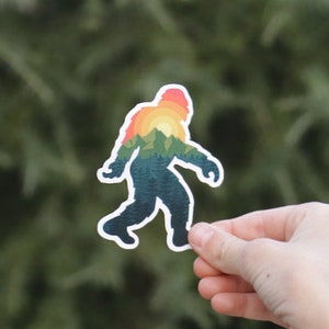 May include: A sticker featuring a silhouette of Bigfoot with a sunset and mountain landscape inside. The sticker is printed on white vinyl and has a glossy finish.