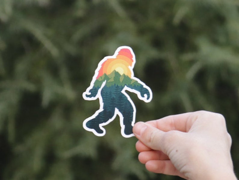 May include: A sticker featuring a silhouette of Bigfoot with a sunset and mountain landscape inside. The sticker is printed on white vinyl and has a glossy finish.