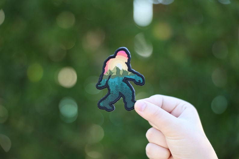Op de afbeelding: Een geborduurde patch met de silhouet van een Bigfoot met een zonsondergang en een berglandschap erin, in tinten groen, zwart en roze.