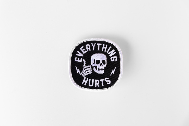 May include: A square embroidered patch with rounded corners featuring a black background and a white border. The design includes a white skull, a skeletal hand giving a thumbs up, two small lightning bolts, and the text EVERYTHING HURTS.