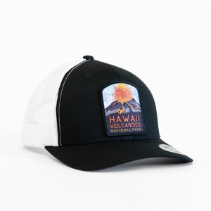 Op de afbeelding: Zwarte en witte truckercap met een patch met een uitbarstende vulkaan en de tekst "HAWAII VOLCANOES NATIONAL PARK".