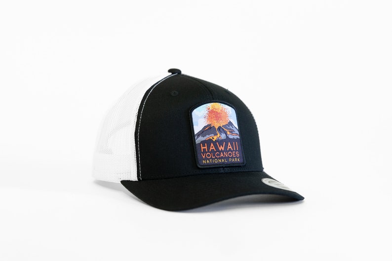 Op de afbeelding: Zwarte en witte truckercap met een patch met een uitbarstende vulkaan en de tekst "HAWAII VOLCANOES NATIONAL PARK".