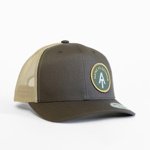 May include: A dark olive green trucker hat with a beige mesh back. The front features a circular patch with a green background, a gold border, and a white Appalachian Trail logo with the text Maine to Georgia printed around the top edge.