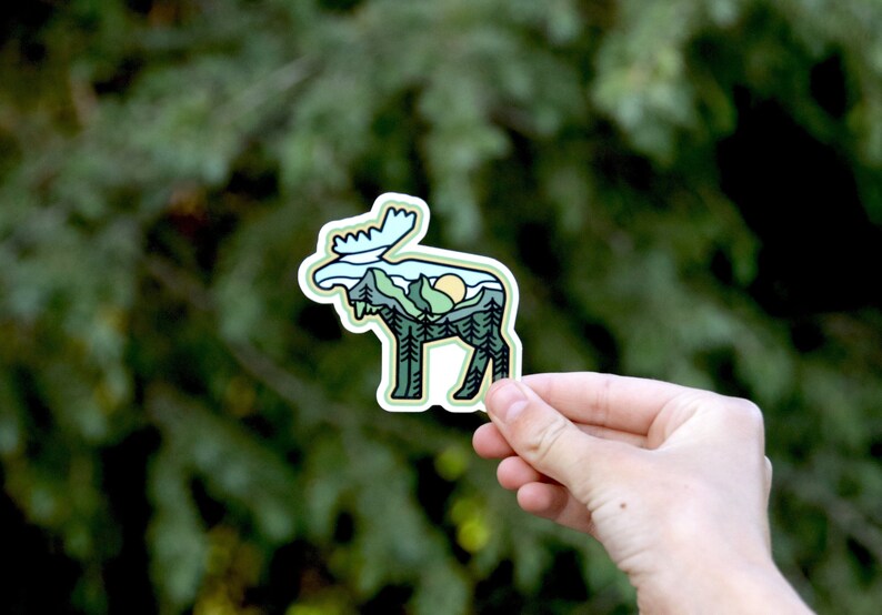 May include: A die-cut sticker featuring a moose silhouette with a landscape design. The design includes mountains, trees, and a sun, all in shades of green, blue, and yellow. The sticker is held against a blurred green background.