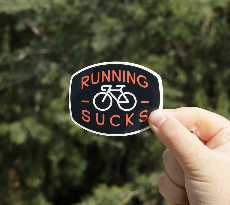 May include: Black and white sticker with the text "Running Sucks" in orange letters. A white bicycle icon is in the centre of the sticker.