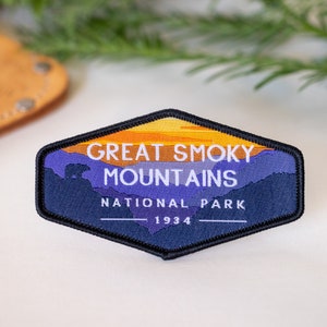 Puede incluir: Un parche bordado azul y naranja con el texto "Great Smoky Mountains National Park 1934". El parche presenta una silueta de monta&ntilde;as y una puesta de sol.