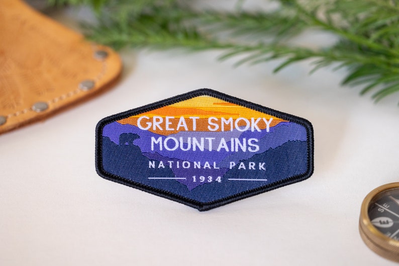 Puede incluir: Un parche bordado azul y naranja con el texto "Great Smoky Mountains National Park 1934". El parche presenta una silueta de monta&ntilde;as y una puesta de sol.