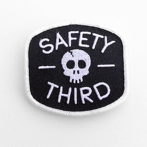 Close on white stitching, the black Safety Third patch shows its skull and white border on a pale surface.