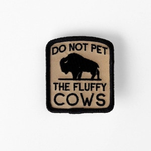 May include: A rectangular embroidered patch with a tan background and a black border. The patch features a black silhouette of a bison in the center, with the black text DO NOT PET THE FLUFFY COWS embroidered above and below the bison graphic.