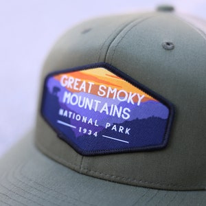 Puede incluir: Gorra de camionero verde y beige con un parche que dice "Great Smoky Mountains National Park 1934". El parche presenta una puesta de sol y monta&ntilde;as.