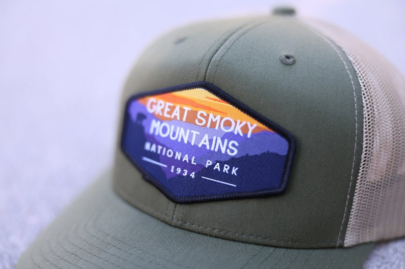 Puede incluir: Gorra de camionero verde y beige con un parche que dice "Great Smoky Mountains National Park 1934". El parche presenta una puesta de sol y monta&ntilde;as.