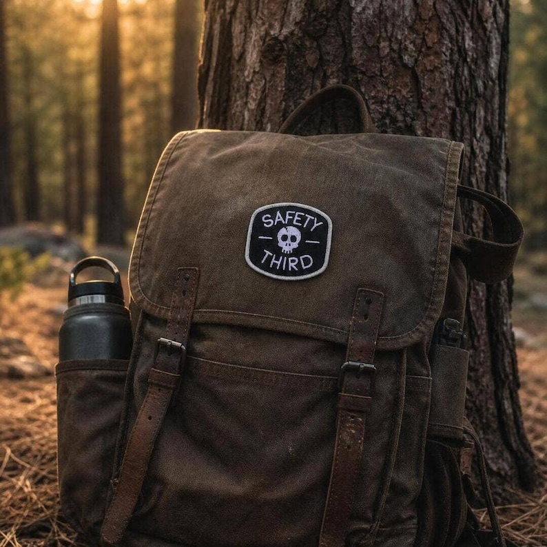 Stitched onto the flap of a waxed canvas backpack, the Safety Third patch rests against a pine trunk at sunset.