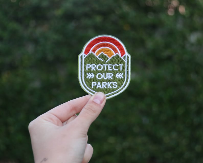 Fingers pinch the lower edge of the Protect Our Parks patch with dense shrubbery behind it.
