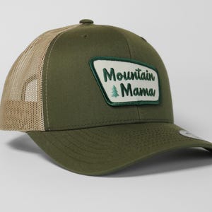 Peut inclure: Casquette de camionneur vert olive avec un dos en filet beige. La casquette présente un écusson rectangulaire avec les mots "Mountain Mama" en blanc et un petit graphisme d'arbre vert.