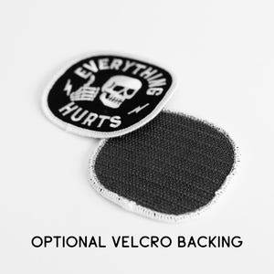May include: A round embroidered patch with a black background and white border. The design features a white skull, a skeletal hand giving a thumbs up, and the text EVERYTHING HURTS. A second patch shows a dark gray hook-and-loop velcro backing.
