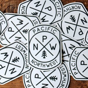A loose pile of Pacific Northwest Compass stickers overlaps across a weathered wooden table.