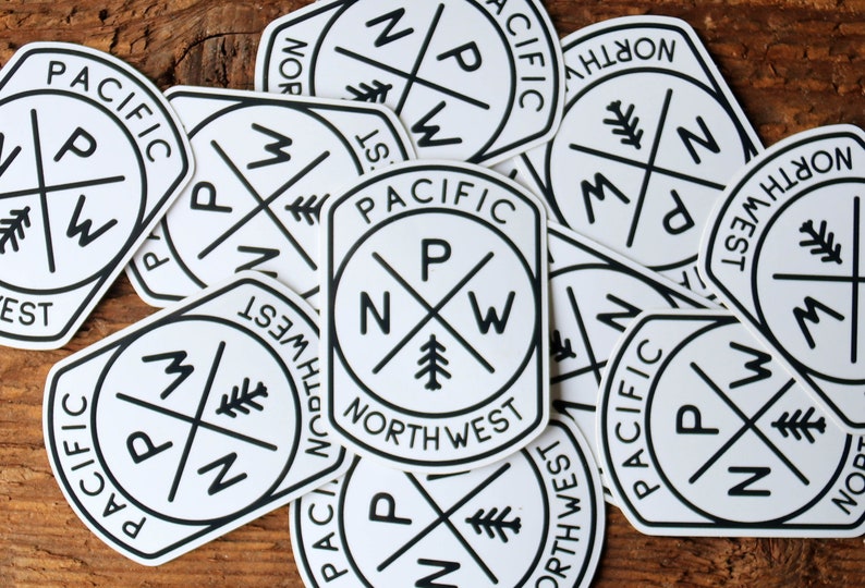 A loose pile of Pacific Northwest Compass stickers overlaps across a weathered wooden table.