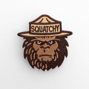 May include: An embroidered patch featuring a brown, furry creature wearing a tan ranger-style hat. The hat has the word SQUATCHY stitched in dark brown letters. The creature has a serious expression with dark brown fur and tan facial details.