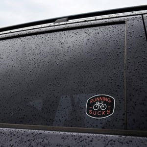 On the rain beaded window of a dark car, the Running Sucks sticker sits low near the door frame.
