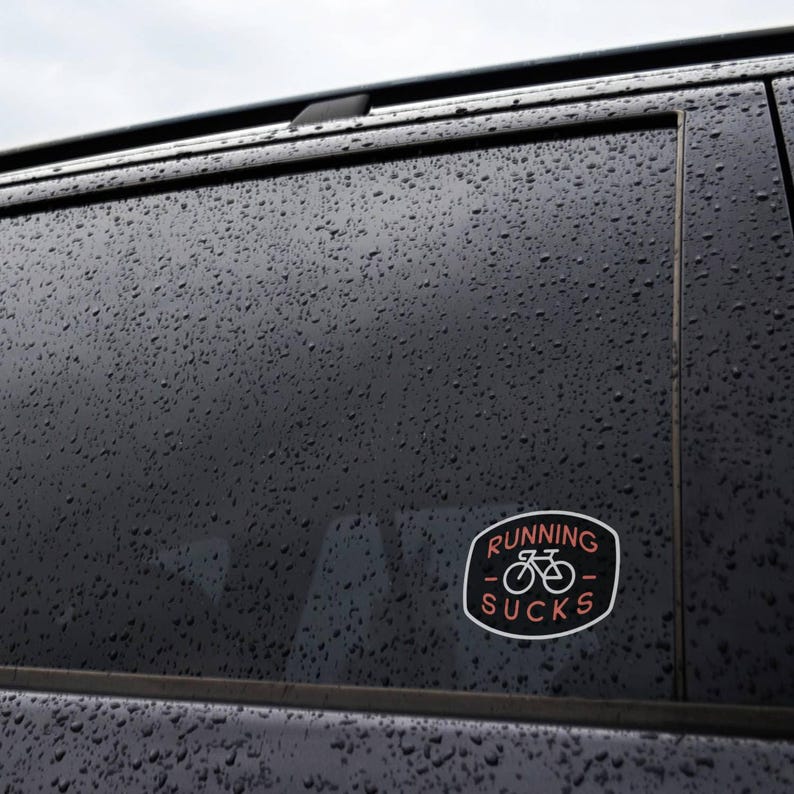 On the rain beaded window of a dark car, the Running Sucks sticker sits low near the door frame.