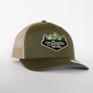 May include: Olive green trucker hat with a tan mesh back. The front features an embroidered patch with green mountains, trees, and the text "The Mountains Are Calling" in white script.