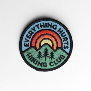 May include: A circular embroidered patch featuring a light blue background with the text "EVERYTHING HURTS" in black. Below, a green mountain range with three black pine trees sits above the text "HIKING CLUB" in black, with a rainbow arch.