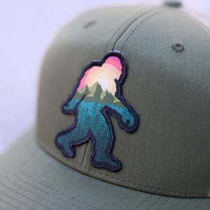 Op de afbeelding: Een groene en beige truckercap met een zwart en kleurrijk patch van een Bigfoot-silhouet met een zonsondergang en bergen op de achtergrond.