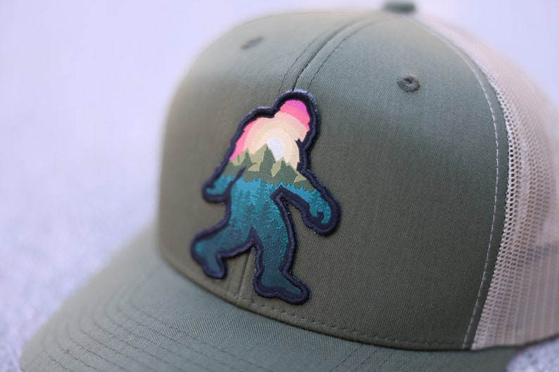 Op de afbeelding: Een groene en beige truckercap met een zwart en kleurrijk patch van een Bigfoot-silhouet met een zonsondergang en bergen op de achtergrond.