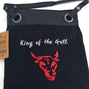 Op de afbeelding: Zwarte canvas schort met een leren bovenkant en verstelbare band. De schort heeft een rode stierenkop afbeelding en de tekst "King of the Grill" geborduurd.