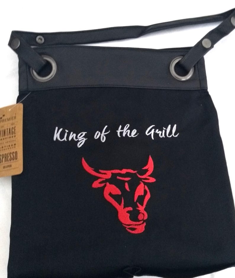 Op de afbeelding: Zwarte canvas schort met een leren bovenkant en verstelbare band. De schort heeft een rode stierenkop afbeelding en de tekst "King of the Grill" geborduurd.