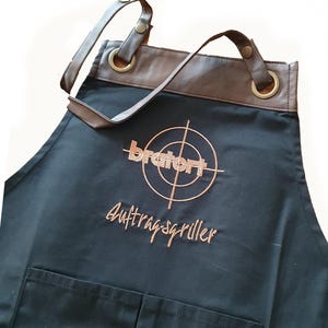 Könnte beinhalten: Schwarze Schürze mit braunem Lederbesatz und verstellbaren Trägern. Die Schürze hat eine große Tasche auf der Vorderseite und ein gesticktes Logo mit der Aufschrift "bratwurst" mit einem Zielgrafik und dem Text "Auftragsgriller".