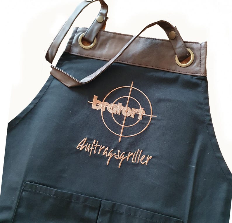 Op de afbeelding: Zwarte schort met bruine leren bies en verstelbare banden. De schort heeft een grote zak aan de voorkant en heeft een geborduurd logo met de tekst "bratwurst" met een doelwitafbeelding en de tekst "Auftragsgriller".